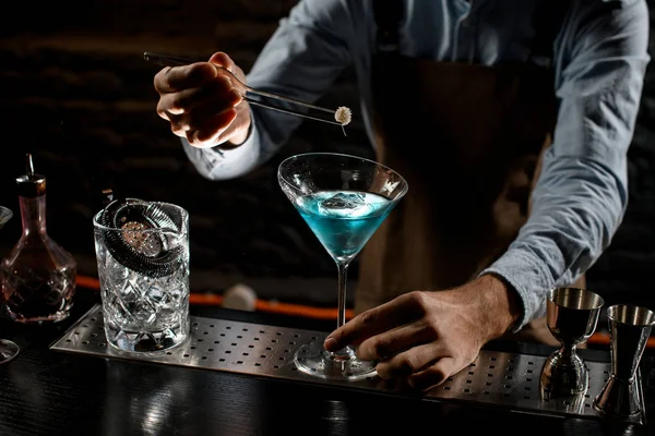 Cantinero profesional que decora un cóctel alcohólico azul en una copa de martini con una pinza de pinzas — Foto de Stock