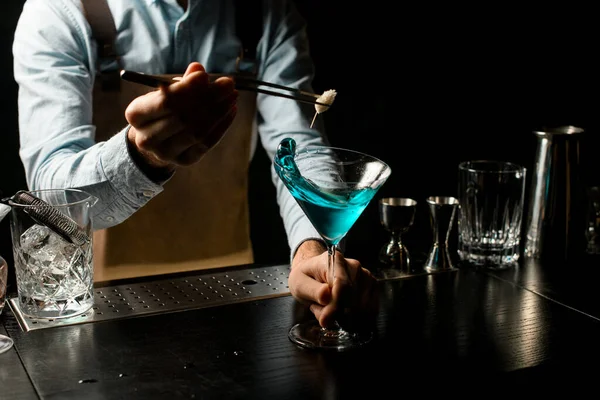 Barista professionista maschile che decora un cocktail alcolico blu in un bicchiere da martini con una spikelet di pinzette — Foto Stock