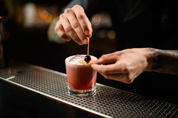 Barman profissional decorando um coquetel alcoólico rosa no copo com uma laranja seca e uma baga no palito — Fotografia de Stock