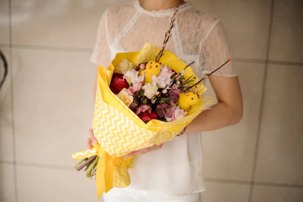 Mulher segurando um buquê de primavera de flores diferentes decoradas com ramos de salgueiro, pequeno brinquedo chiken envolto em papel amarelo — Fotografia de Stock