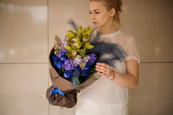 Chica sosteniendo una composición de ramo de flores de primavera oscura con flores, plumas azules en un papel de oro — Foto de Stock