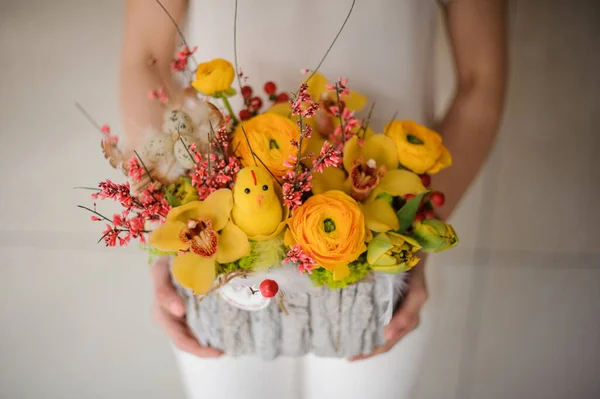 Woman holding a composition in a wood trunk piece with yellow flowers, branches and toy chicken — Stockfoto