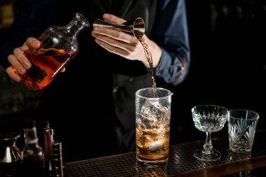 Barman measures the ingredients for cocktail from bottle with brown alcohol drink.