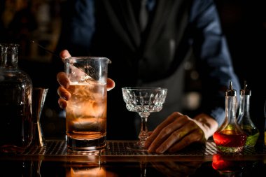 On the bar stands a glass of cocktail with ice and a wineglass.