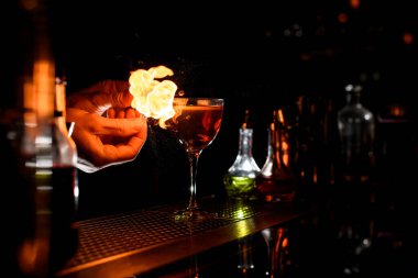 close-up bartenders hands which sprinkling of cocktail glass by juice of citrus and sets fire.