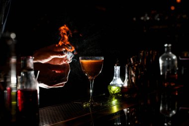 magnificent close-up of bartenders hands lighting a flame near glass with cocktail.