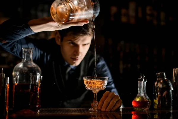 Young bartender pours a ready-made cold cocktail into wineglass and looks at it — Stockfoto