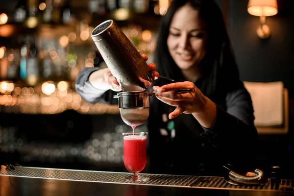 Mulher sorridente bartender derrama coquetel de shaker em vidro usando peneira . — Fotografia de Stock