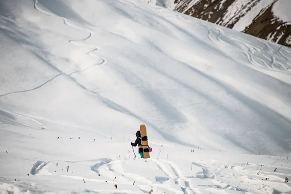 Homme avec snowboard sur le dos se tient devant une énorme montagne enneigée . — Photo