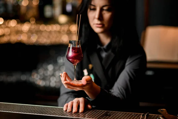 Brünette Frau Barkeeperin hält Weinglas mit rotem Cocktail, der von zwei Stöcken verziert wird. — Stockfoto