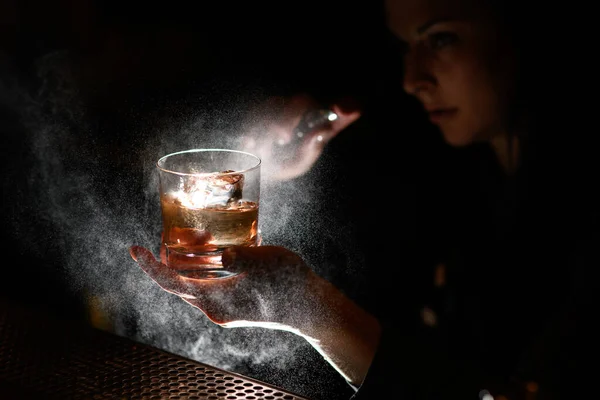 En barra oscura mujer sostiene a mano vaso con bebida y spray sobre ella . — Foto de Stock