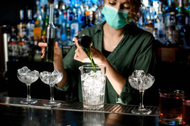 Tıbbi maskeli kız barmen bardağa buzlu sıvı döker.
