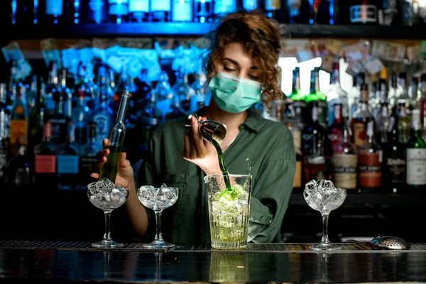 Girl bartender in medical mask pours green liquid into glass with ice using beaker. — Stock Photo, Image