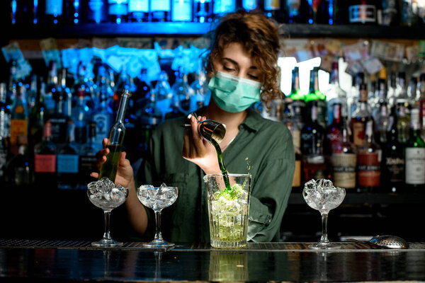 girl bartender in medical mask pours green liquid into glass with ice using beaker.