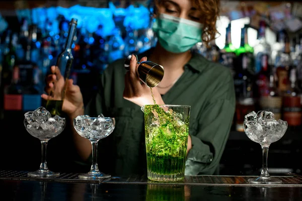 Close-up girl bartender in medical mask carefully pours green liquid into glass with ice using beaker. — Stock Photo, Image