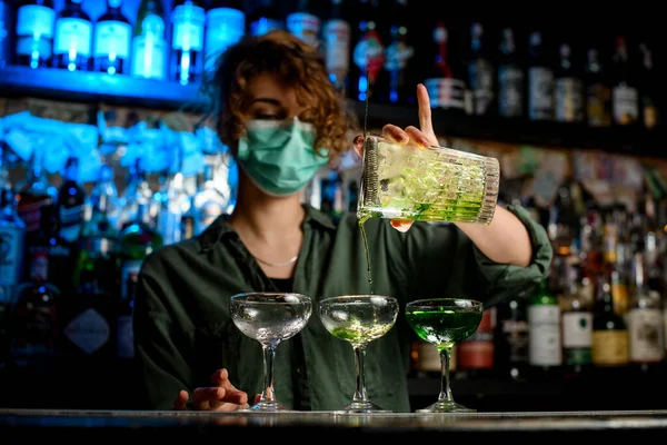Woman bartender in medical mask carefully pours drink from large glass into wineglasses. — Stock Photo, Image