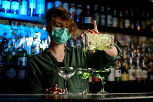 Young woman bartender in medical mask carefully pours drink from large glass into wineglasses. — Stock Photo, Image