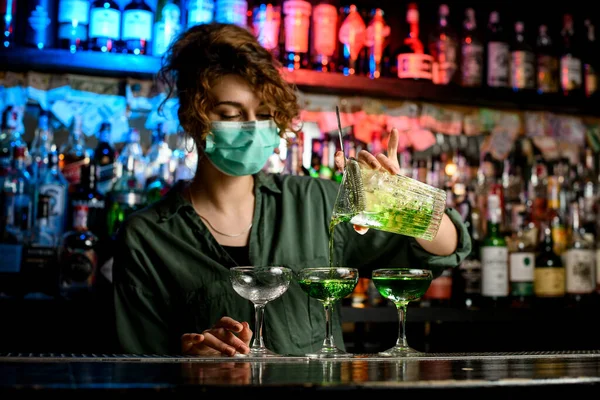 Νεαρή γυναίκα μπάρμαν σε ιατρική μάσκα επαγγελματικά χύνει ποτό από μεγάλο ποτήρι σε ποτήρια κρασιού. — Φωτογραφία Αρχείου