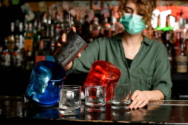 Camarera chica en máscara médica comenzar a verter bebida de vidrio de acero en vasos con hielo . — Foto de Stock