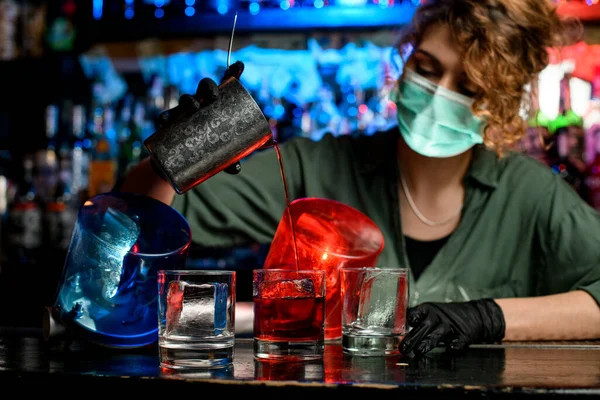 Barmanka v lékařské masce a černé rukavice přesné nalévání nápoje z ocelového skla do sklenic s ledem. — Stock fotografie