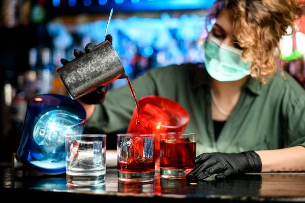 Chica camarera joven en máscara médica y guantes negros precisa verter bebida de vidrio de acero en vasos con hielo . — Foto de Stock