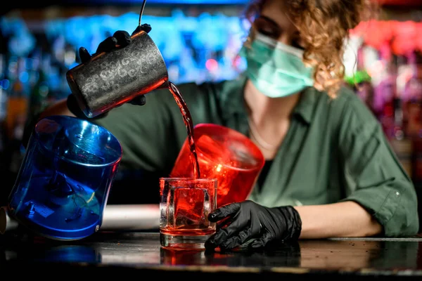 Young bartender girl in medical mask and black gloves professionally pour drink from steel glass into glasses with ice.