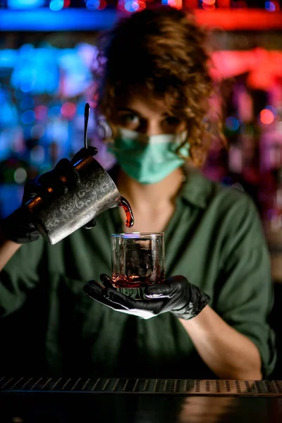 Mujer barman en máscara médica y guantes negros sostiene el vidrio y vierte energía bebida en ella . — Foto de Stock