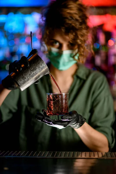Mujer barman en máscara médica y guantes negros sostiene el vidrio y vierte suavemente beber en ella . — Foto de Stock