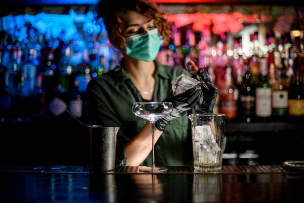 Mujer barman en máscara médica y guantes negros divide trozos de hielo para hacer cóctel . — Foto de Stock
