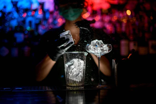 Primer plano. Mujer barman en máscara médica y guantes negros con dispositivo especial precisa rompe pedazos de hielo . — Foto de Stock