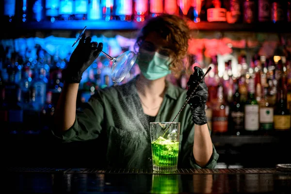 Jovem no balcão bar em máscara médica e luvas pretas precisas detém vidro e spray para ele . — Fotografia de Stock