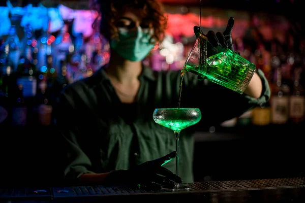 Νεαρή μπαργούμαν με ιατρική μάσκα και μαύρα γάντια με επαγγελματισμό χύστε πράσινο ποτό από γυαλί στο κρασί. — Φωτογραφία Αρχείου