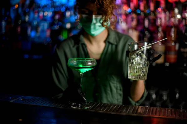 Chica joven en el mostrador de bar en máscara médica y guantes negros sostiene vidrio con cóctel verde . — Foto de Stock