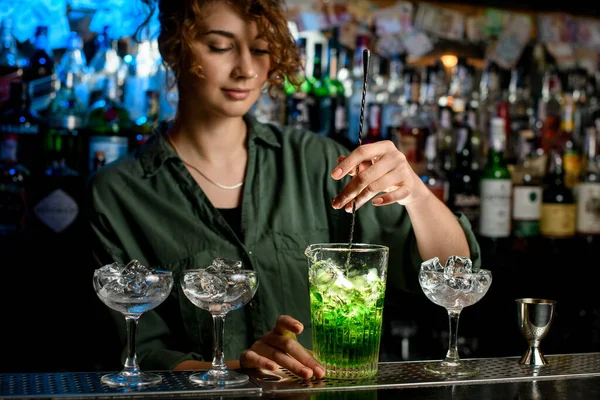 Young woman bartender actively mixes cocktail in glass with ice.