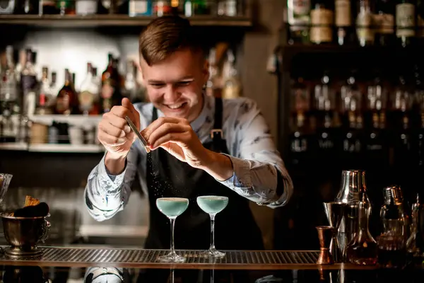 Ung leende bartender dekorerar två glas med cocktails. — Stockfoto