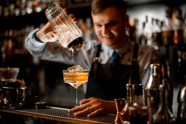 Усміхнений молодий бармен наливає готовий коктейль зі скляного шейкера в склянку з льодом . — стокове фото