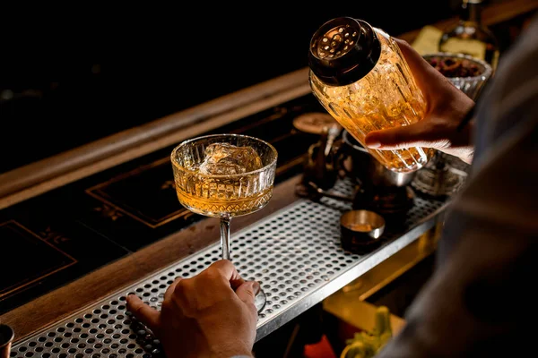Barman nalévá hotový koktejl ze skleněného třepače do skla s ledem na barovém pultu. — Stock fotografie
