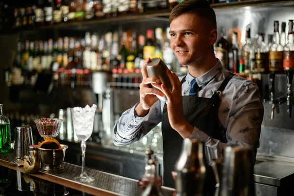Jovem garçom sorrindo em pé no balcão bar com agitador em suas mãos . — Fotografia de Stock