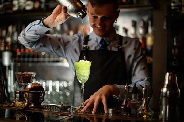 Mladý usměvavý barman nalévá koktejl z třepačky do skla s ledem. — Stock fotografie
