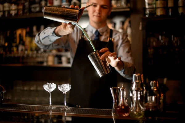 Jovem barman derrama coquetel em shaker. Dois copos com suporte de gelo no bar . — Fotografia de Stock