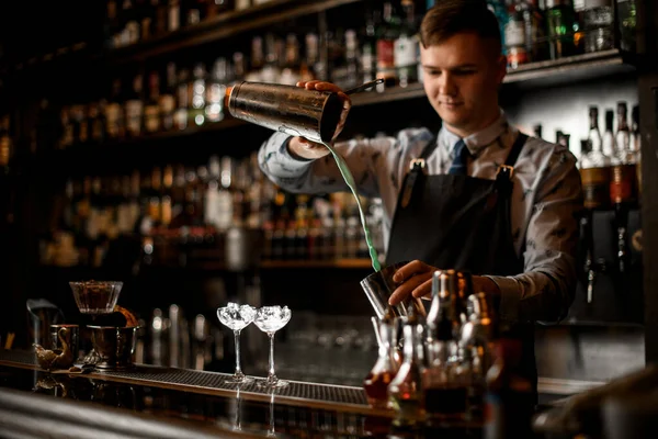 Junge gut aussehende Barkeeper gießt Cocktail in Shaker. Zwei Gläser mit Eis stehen auf der Theke. — Stockfoto