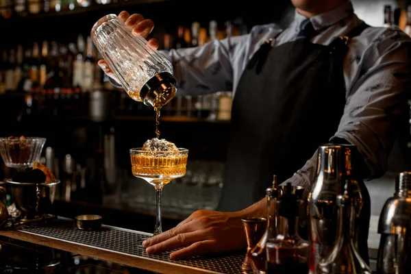 Professional bartender in black apron pours drink from shaker into glass. — Stock Photo, Image