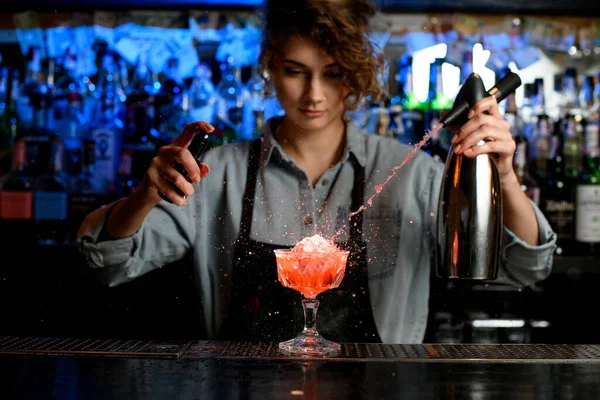 Vrouw barman expressieve gieten cocktail van staal sifon naar glas op bar teller. — Stockfoto