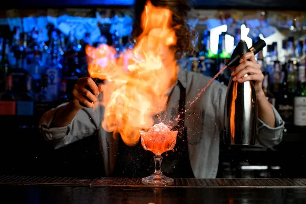 Женщина-бармен, распыляющая огонь вокруг коктейльного стекла на барной стойке — стоковое фото