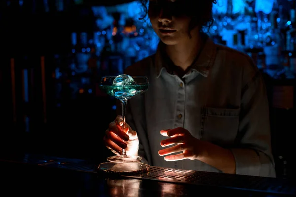 lady at bar carefully holds beautiful glass of blue cocktail.