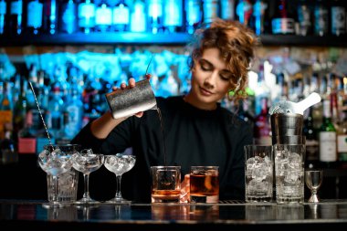 Young woman barman preparing cocktail and carefully pouring it into glass. clipart
