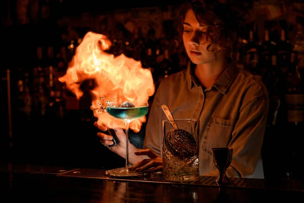 Женщина в темном баре профессионально разводит огонь над бокалом вина с коктейлем . — стоковое фото
