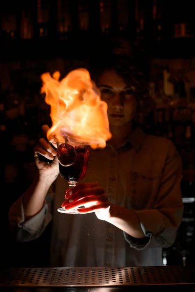 Девушка в темном баре держит бокал с коктейлем и разжигает над ним огонь. . — стоковое фото
