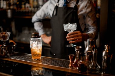 Barmen bardağı buzla tutuyor. Cam kokteyl karıştırıcı tezgahın üzerinde duruyor..