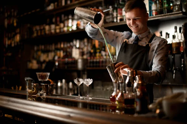 Young smiling barman professionally pours cocktail in shaker at bar.